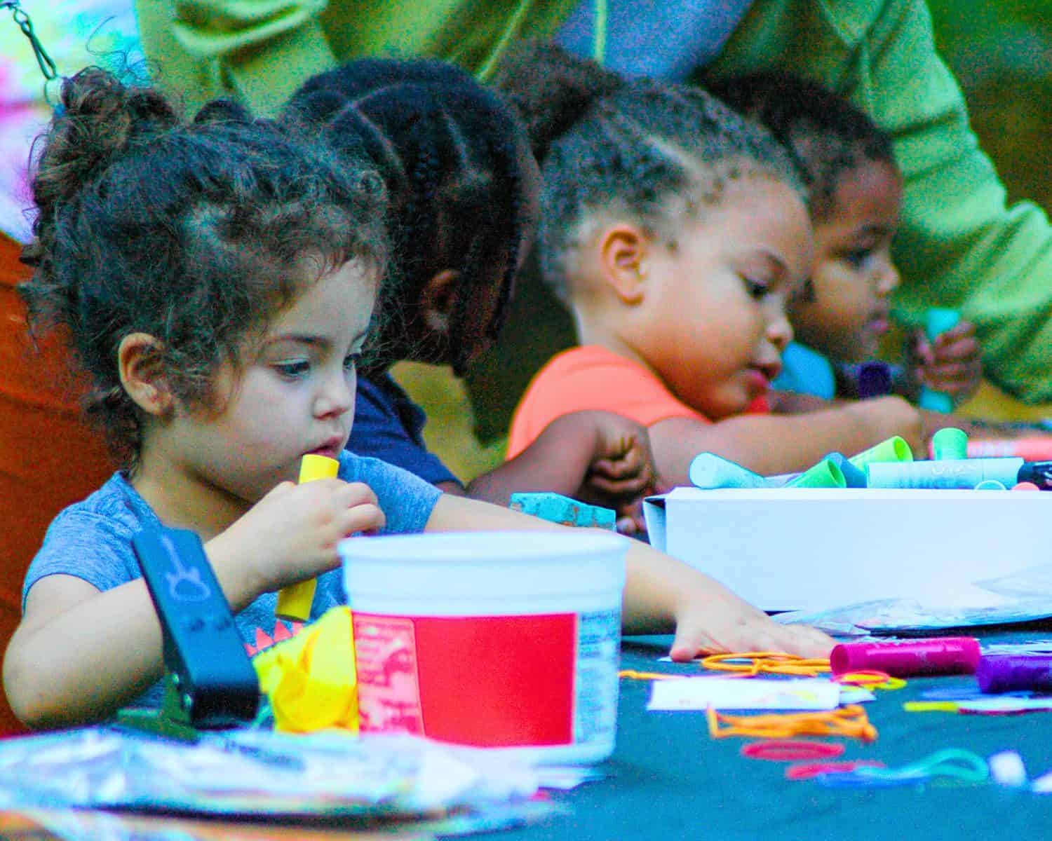 Arts-in-Healing-Website-Page-2-1-web A close-up, candid photo of four toddlers, from a diverse group, intensely focused on a table craft activity during an outdoor community event.